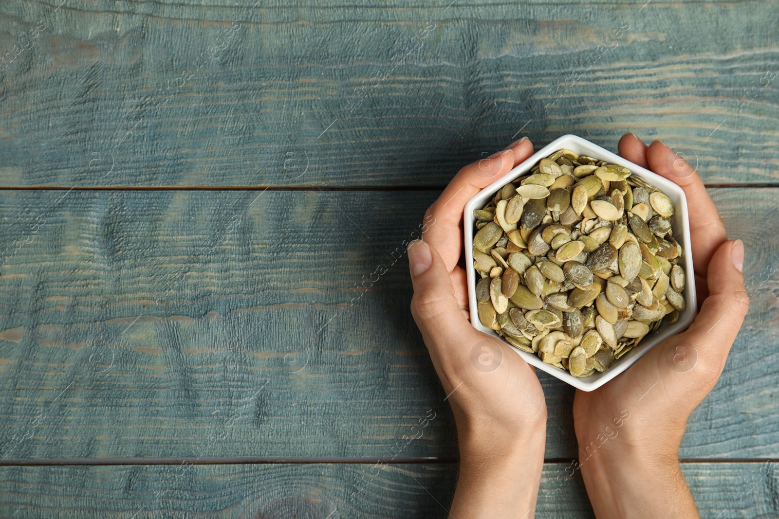 Photo of Young woman with bowl of raw pumpkin seeds at blue wooden table, top view. Space for text