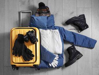 Photo of Suitcase with warm clothes and camera on wooden floor, flat lay. Winter vacation