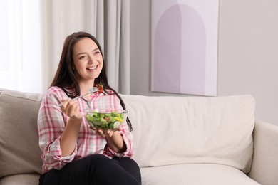 Photo of Beautiful overweight woman eating salad in living room, space for text. Healthy diet