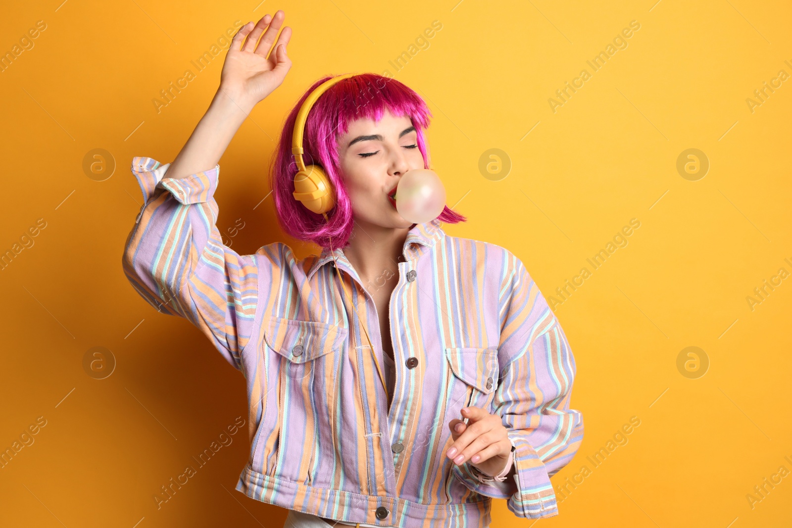 Photo of Fashionable young woman in colorful wig with headphones blowing bubblegum on yellow background, space for text