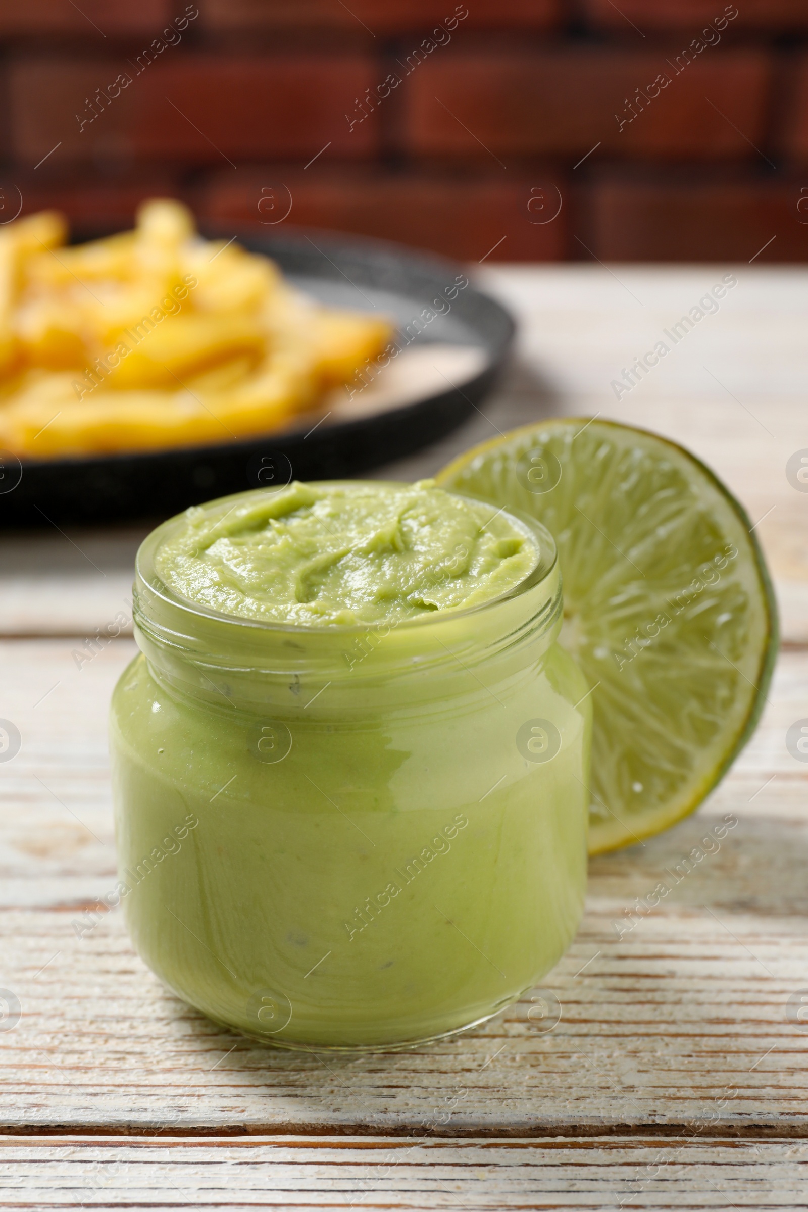 Photo of Jar of avocado dip, lime and plate with french fries on white wooden table