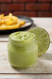 Photo of Jar of avocado dip, lime and plate with french fries on white wooden table