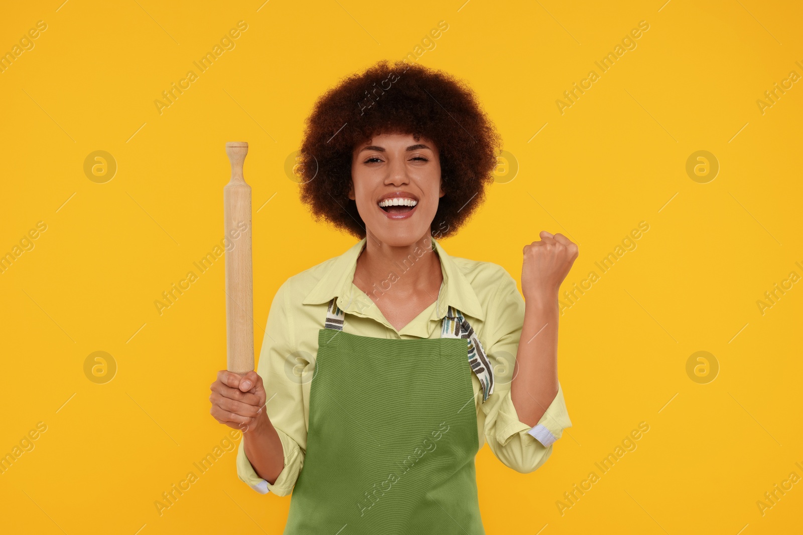 Photo of Emotional young woman in apron holding rolling pin on orange background