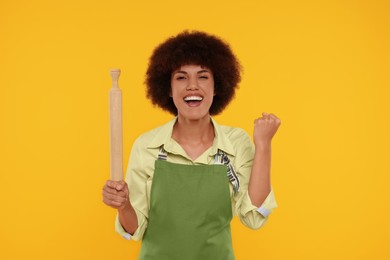 Emotional young woman in apron holding rolling pin on orange background