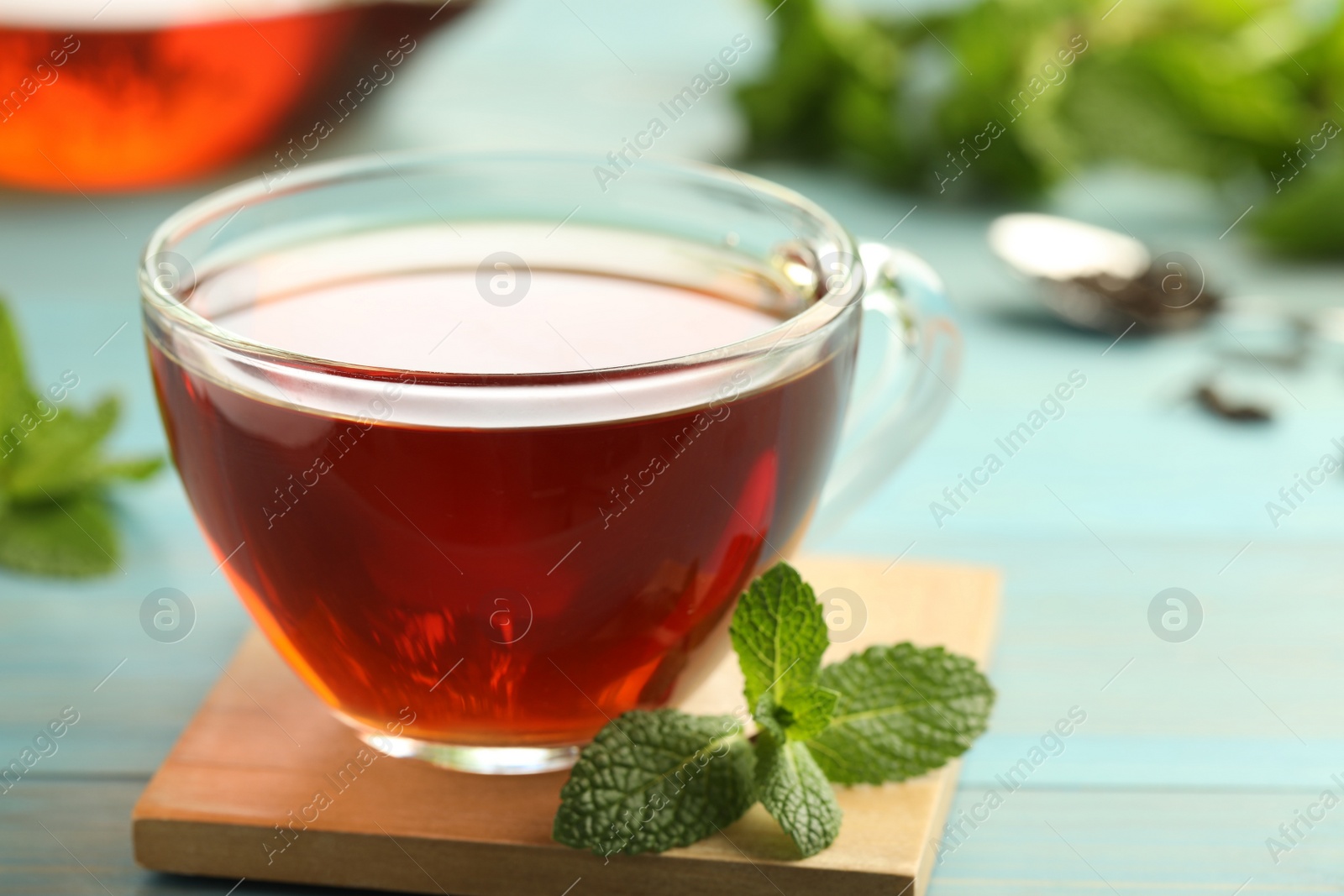 Photo of Glass cup of aromatic black tea with fresh mint on light blue wooden table against blurred background, closeup
