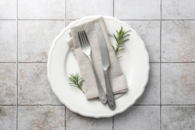 Photo of Stylish setting with cutlery, napkin, rosemary and plate on light tiled table, top view