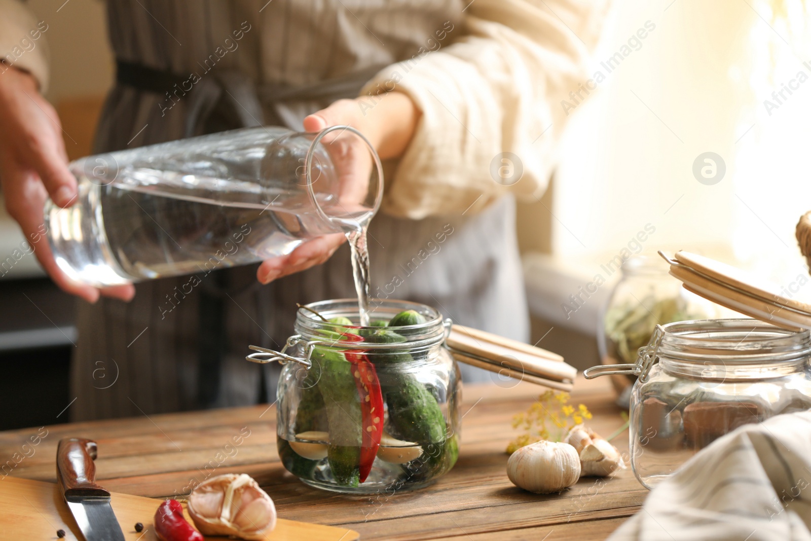 Photo of Woman pouring water into jar with cucumbers at wooden table, closeup. Pickling vegetables