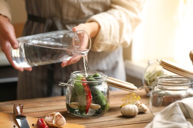 Photo of Woman pouring water into jar with cucumbers at wooden table, closeup. Pickling vegetables