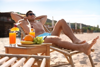 Photo of Happy man resting on sunny beach at resort, focus on table with drinks and fruits
