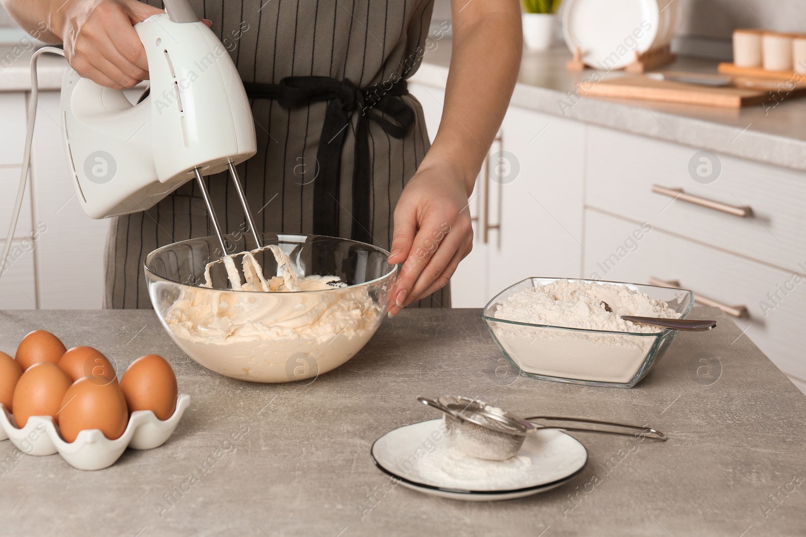 Photo of Woman whipping white cream with mixer at light grey table in kitchen, closeup