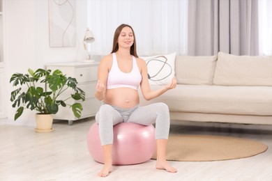 Photo of Pregnant woman meditating on fitness ball in room. Home yoga