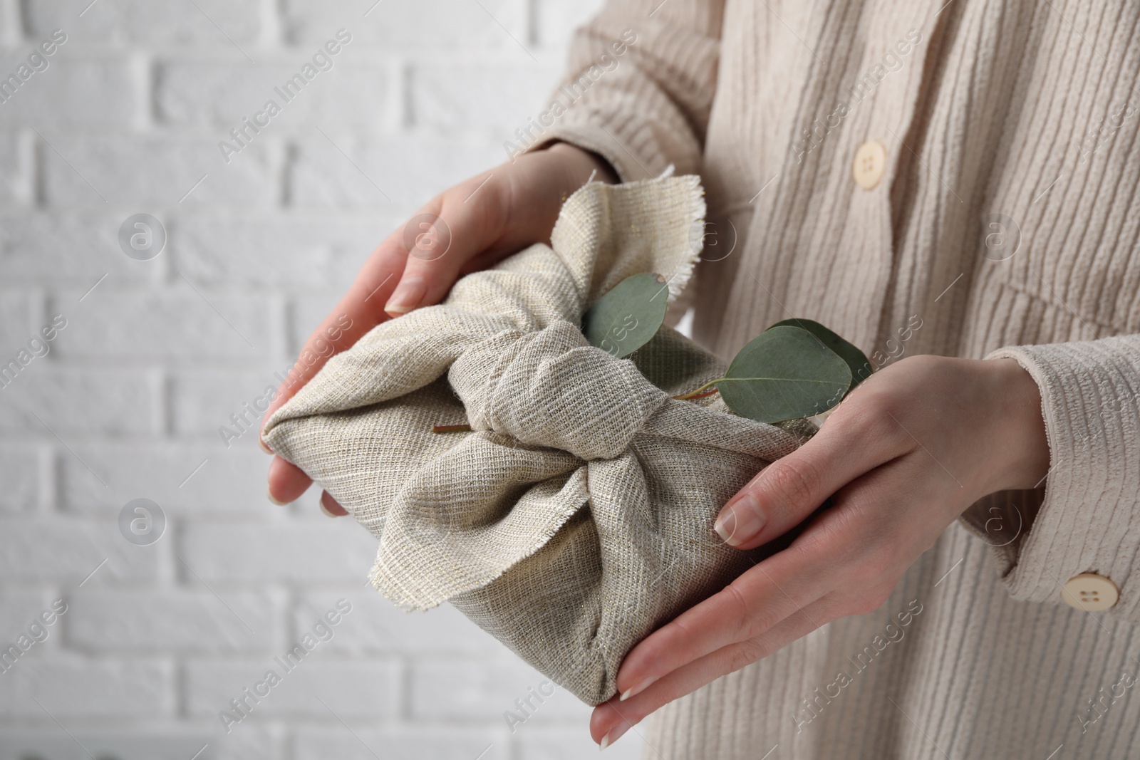 Photo of Furoshiki technique. Woman holding gift packed in fabric and decorated with eucalyptus branch against white wall, closeup