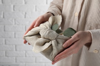 Photo of Furoshiki technique. Woman holding gift packed in fabric and decorated with eucalyptus branch against white wall, closeup