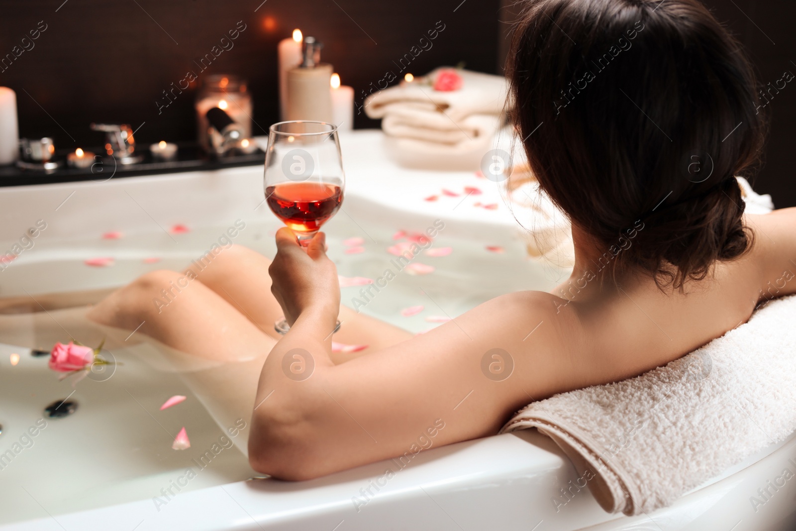 Photo of Woman holding glass of wine while taking bath with rose petals, back view. Romantic atmosphere