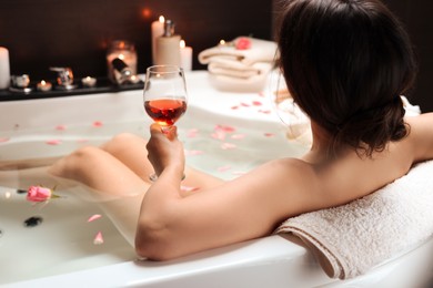Woman holding glass of wine while taking bath with rose petals, back view. Romantic atmosphere