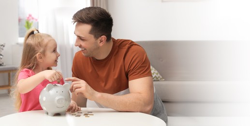 Father and daughter putting coin into piggy bank at table indoors, space for text. Banner design