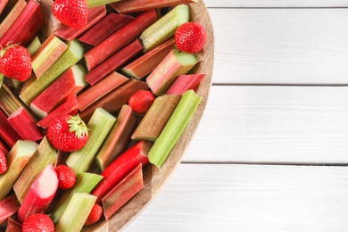 Photo of Cut fresh rhubarb stalks and strawberries on white wooden table, top view. Space for text