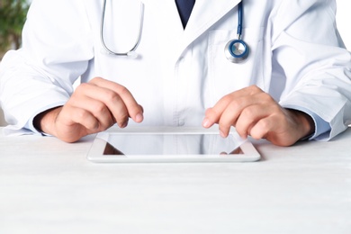 Male doctor with modern tablet sitting at table indoors, closeup