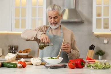 Man pouring oil into bowl with fresh salad at table in kitchen