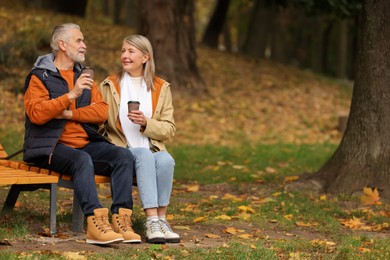 Affectionate senior couple with cups of coffee on wooden bench in autumn park, space for text