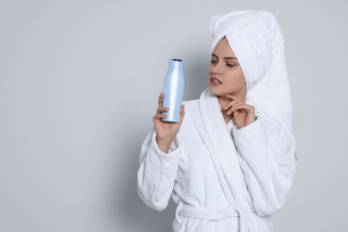 Photo of Beautiful young woman in bathrobe holding bottle of shampoo on light grey background. Space for text