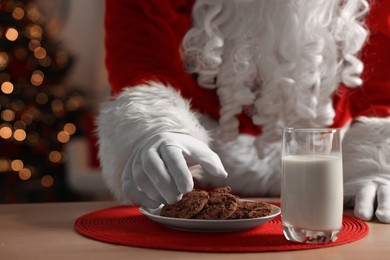 Photo of Santa Claus taking cookies from plate on table in room, closeup. Merry Christmas