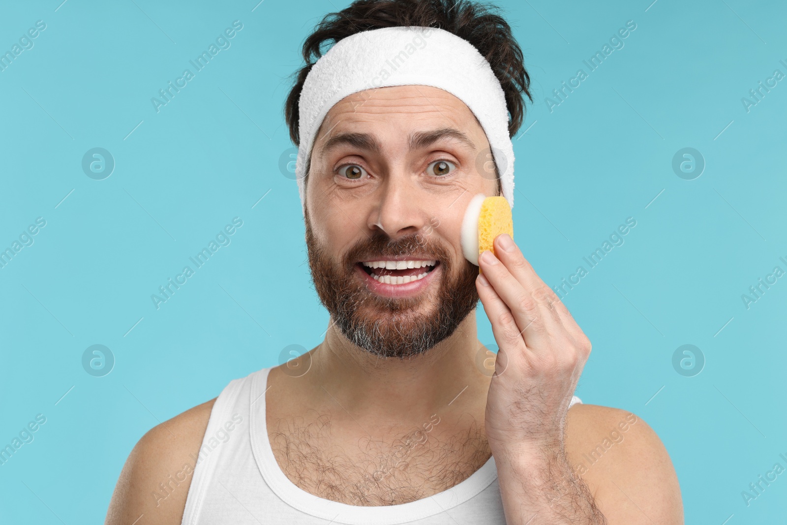 Photo of Man with headband washing his face using sponge on light blue background