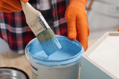 Photo of Woman dipping brush into bucket of light blue paint at wooden table indoors, closeup