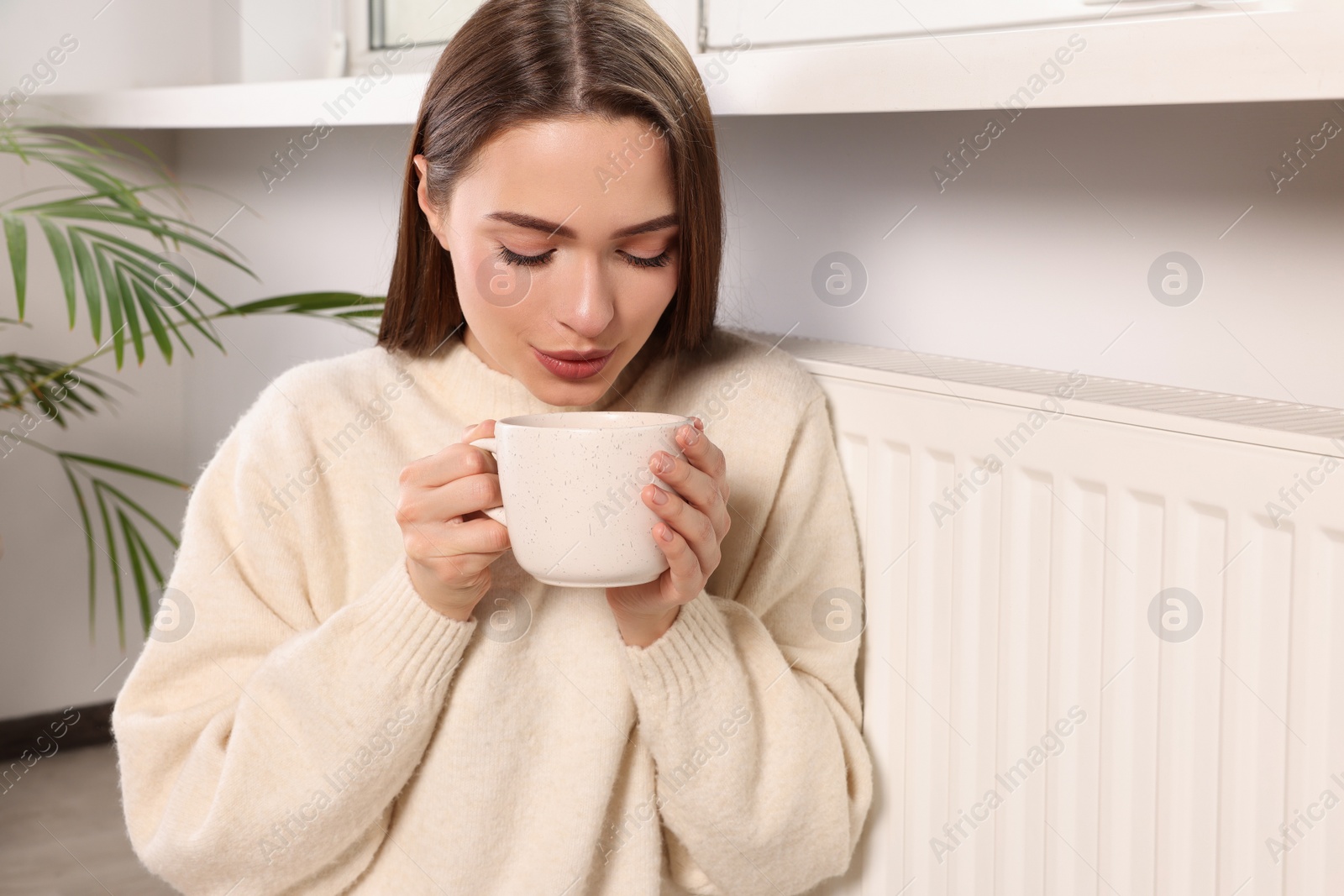 Photo of Woman holding cup with hot drink near heating radiator indoors