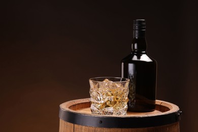 Whiskey with ice cubes in glass and bottle on wooden barrel against dark background, space for text