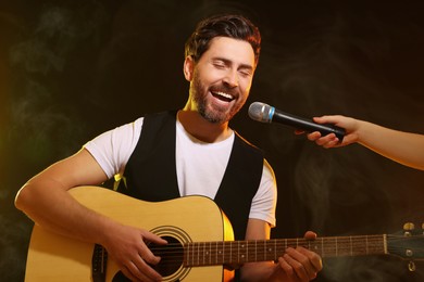 Image of Handsome man with acoustic guitar singing while woman holding microphone on stage in color lighted smoke