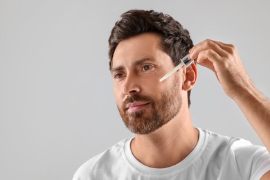 Handsome man applying cosmetic serum onto his face on light grey background. Space for text