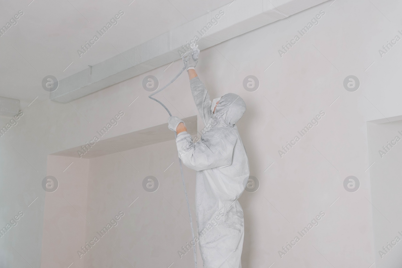 Photo of Decorator in protective overalls painting ceiling with spray gun indoors