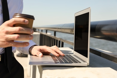 Young man with paper cup of coffee working on laptop outdoors, closeup