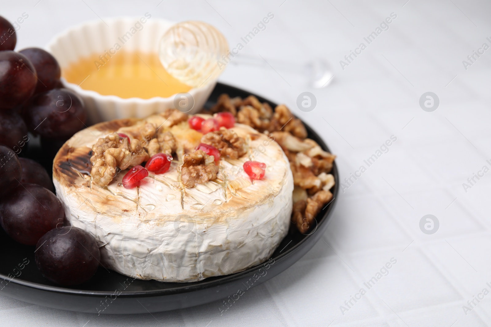 Photo of Plate with tasty baked camembert, honey, grapes, walnuts and pomegranate seeds on white tiled table, closeup. Space for text