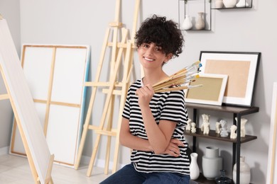 Young woman holding brushes near easel with canvas in studio