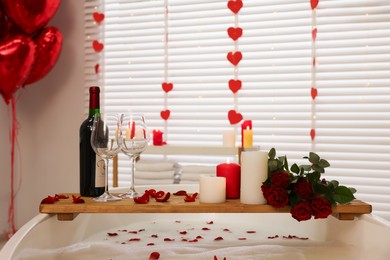 Photo of Wooden tray with wine, burning candles and rose petals on tub in bathroom. Valentine's day celebration