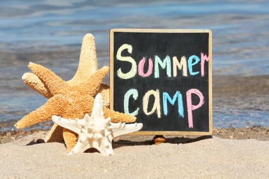 Photo of Little blackboard with text SUMMER CAMP and sea stars on sand near sea