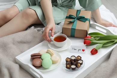 Photo of Tasty breakfast served in bed. Woman with tea, desserts, gift box, flowers and I Love You card at home, closeup