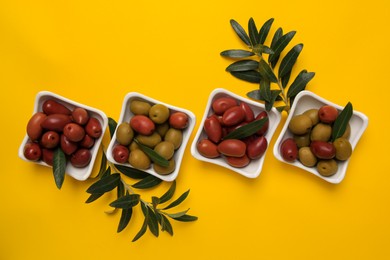 Different fresh olives and green leaves on yellow background, flat lay