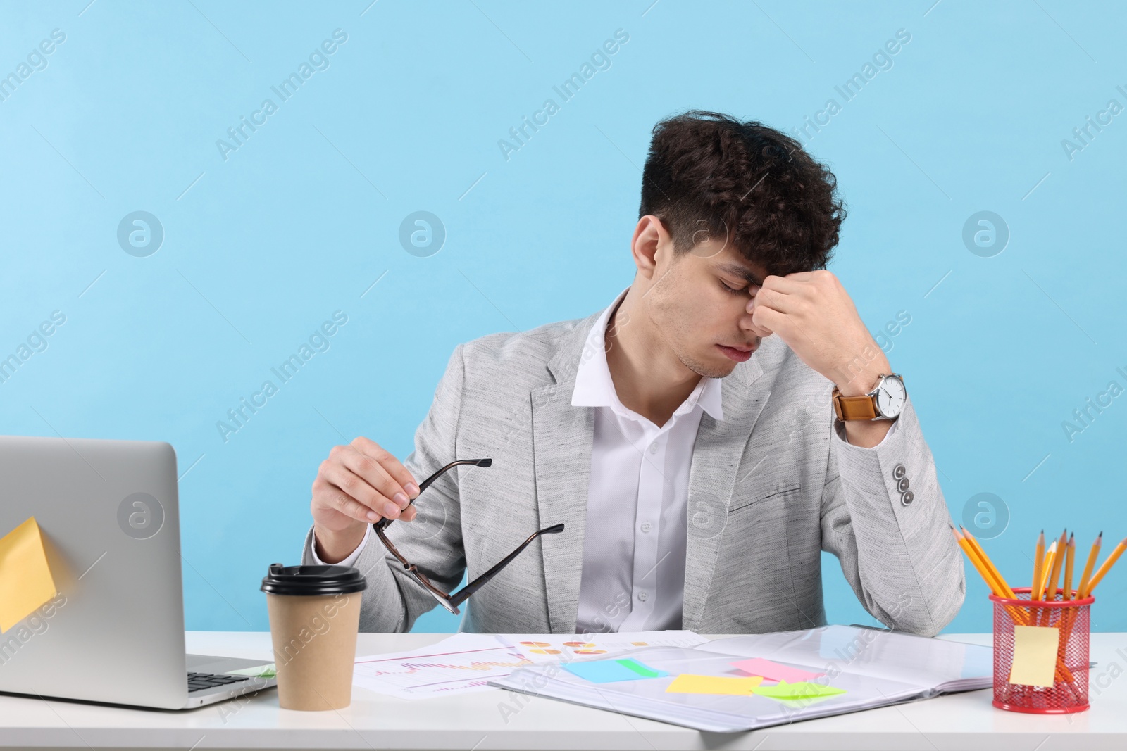 Photo of Tired young man working at white table on light blue background. Deadline concept