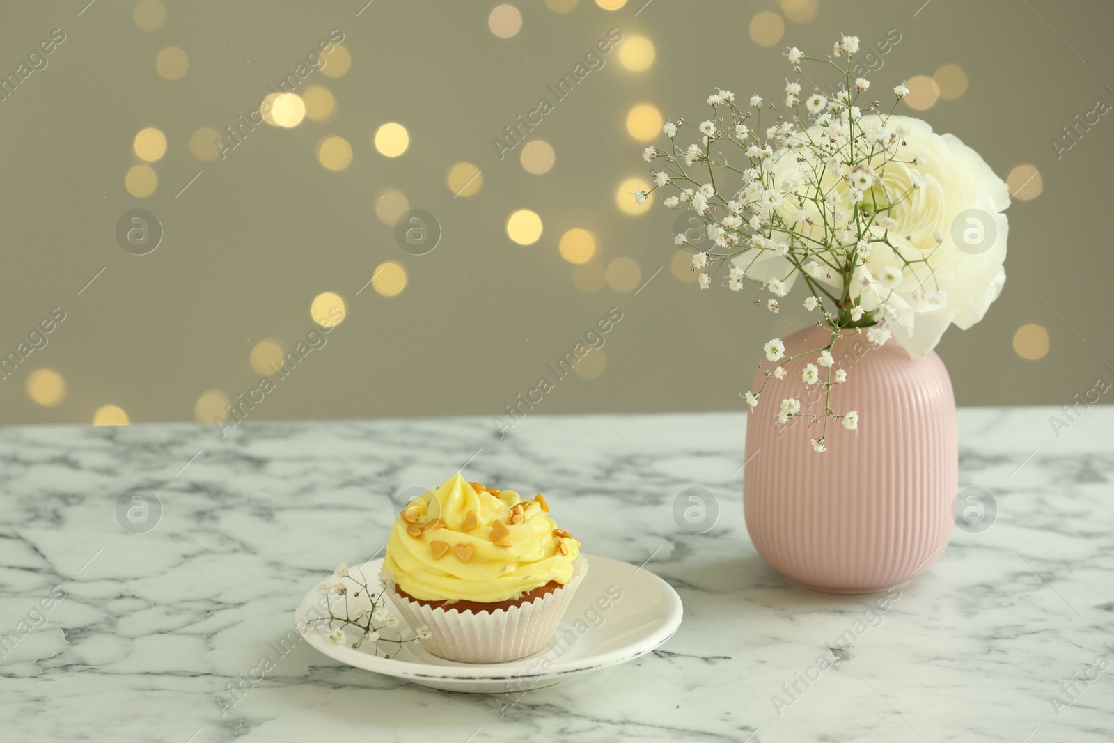 Photo of Delicious cupcake with yellow cream and flowers on white marble table against blurred lights