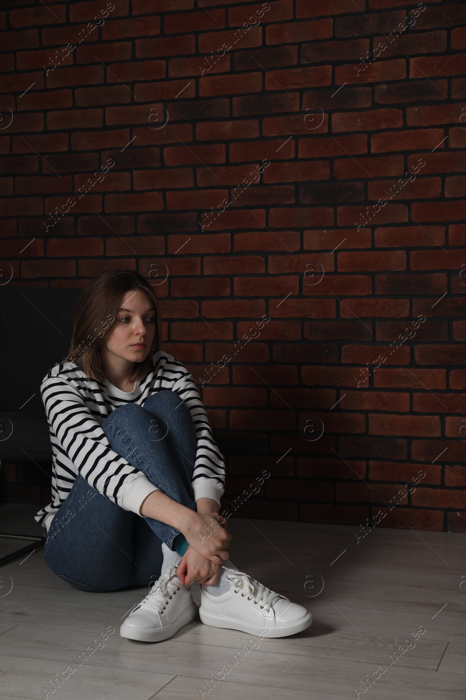 Photo of Sad young woman sitting on floor near brick wall indoors