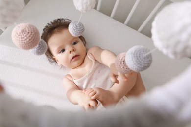 Photo of Cute little baby lying in crib with hanging mobile, top view