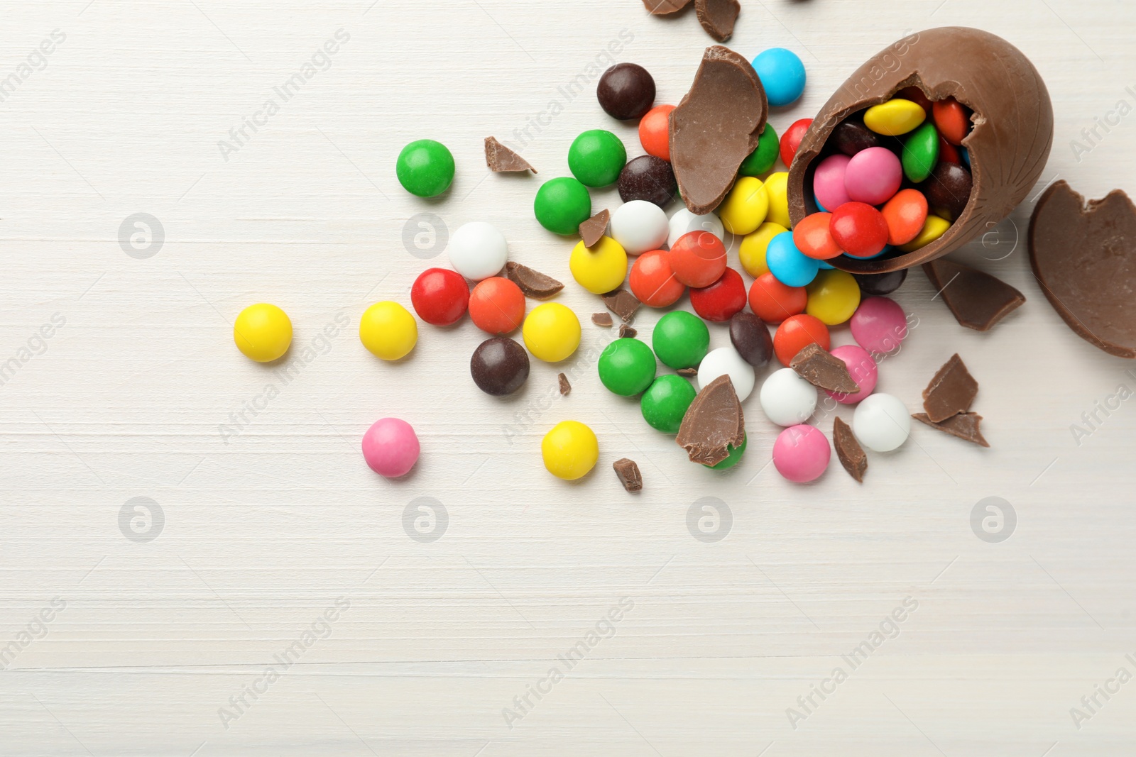 Photo of Broken chocolate egg and colorful candies on white wooden table, flat lay. Space for text