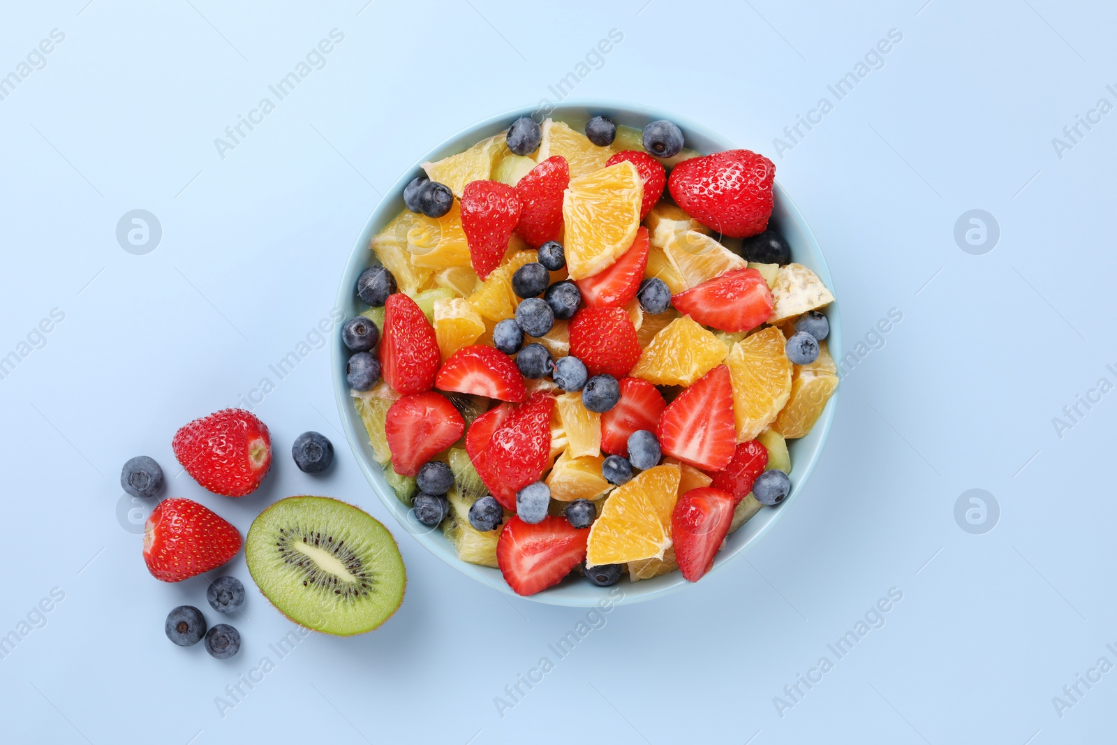 Photo of Yummy fruit salad in bowl and ingredients on light blue background, flat lay