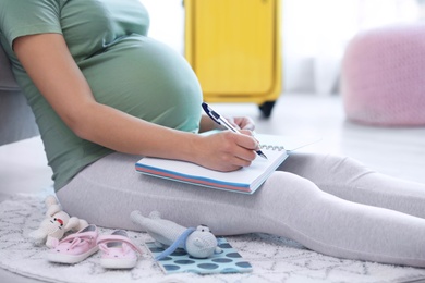 Pregnant woman making list while packing suitcase for maternity hospital at home, closeup