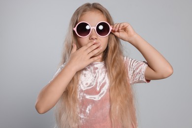 Girl in stylish sunglasses covering mouth with hand on light grey background