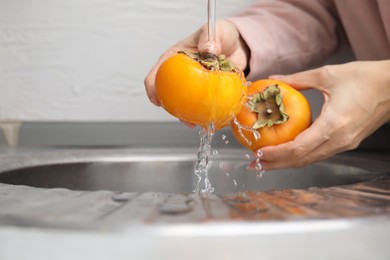 Photo of Woman washing delicious ripe juicy persimmons under tap water indoors, closeup