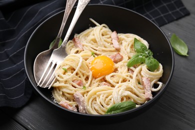 Photo of Bowl of delicious pasta Carbonara with egg yolk and cutlery on black wooden table, closeup